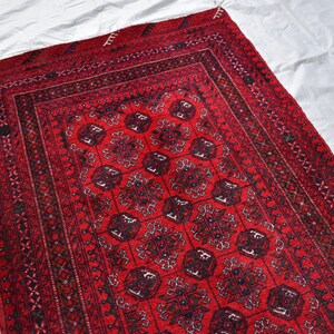 May include: A rectangular red rug with a detailed geometric pattern. The rug features a central field of repeating floral motifs in black and dark red, framed by multiple borders with intricate designs. The rug is placed on a white surface.