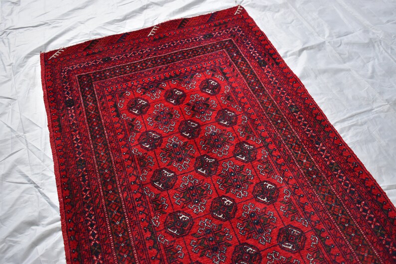 May include: A rectangular red rug with a detailed geometric pattern. The rug features a central field of repeating floral motifs in black and dark red, framed by multiple borders with intricate designs. The rug is placed on a white surface.