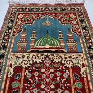 May include: A traditional prayer rug with a blue background and intricate floral designs. The rug features a mosque with a green dome and minarets, surrounded by a red and gold border.