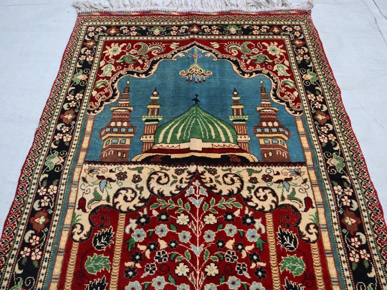 May include: A traditional prayer rug with a blue background and intricate floral designs. The rug features a mosque with a green dome and minarets, surrounded by a red and gold border.