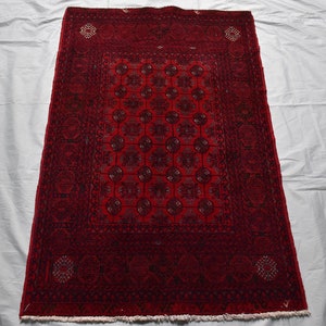 May include: A rectangular, hand-woven rug with a deep red field and intricate geometric patterns. The rug features a central design of repeating circular motifs in shades of black and dark red, framed by a detailed border. The rug's dimensions are not visible.
