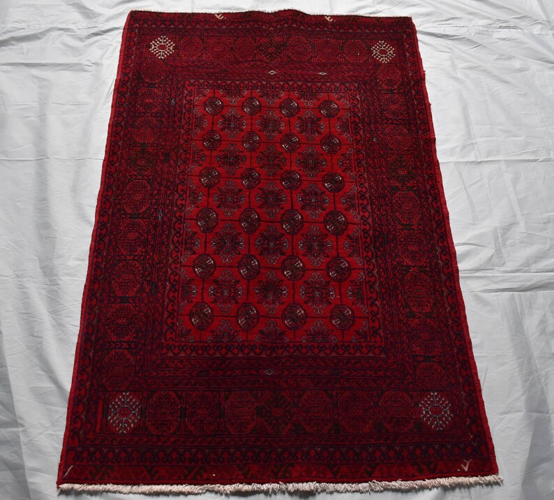 May include: A rectangular, hand-woven rug with a deep red field and intricate geometric patterns. The rug features a central design of repeating circular motifs in shades of black and dark red, framed by a detailed border. The rug's dimensions are not visible.