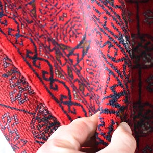 May include: Close-up of a folded red rug with intricate geometric patterns. The rug features a deep red background with dark blue and brown accents. The design includes repeating shapes and borders.