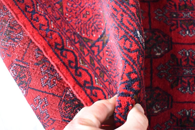 May include: Close-up of a folded red rug with intricate geometric patterns. The rug features a deep red background with dark blue and brown accents. The design includes repeating shapes and borders.