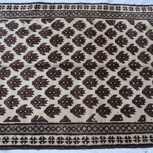 May include: A cream-colored rug with a brown geometric pattern. The rug has a border with a repeating pattern of brown and cream.