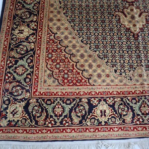Puede incluir: Una alfombra azul marino con un patr&oacute;n floral beige y rojo. La alfombra tiene un borde de flecos.