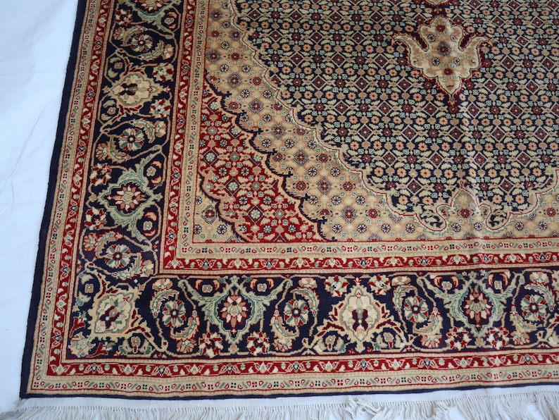 Puede incluir: Una alfombra azul marino con un patr&oacute;n floral beige y rojo. La alfombra tiene un borde de flecos.