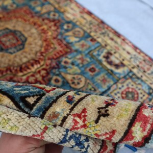 May include: A rolled-up, intricately patterned rug with a central medallion design. The rug features a palette of beige, red, blue, and touches of green. The detailed patterns and colors suggest a traditional, handcrafted style.