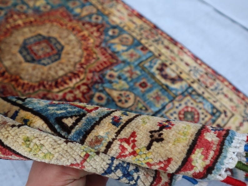 May include: A rolled-up, intricately patterned rug with a central medallion design. The rug features a palette of beige, red, blue, and touches of green. The detailed patterns and colors suggest a traditional, handcrafted style.