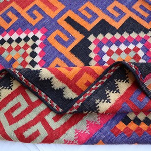 May include: A vibrant, geometric patterned rug with a folded edge. The rug features a mix of colours including red, orange, purple, black, and pink. The design includes repeating geometric shapes and patterns, creating a visually striking textile.