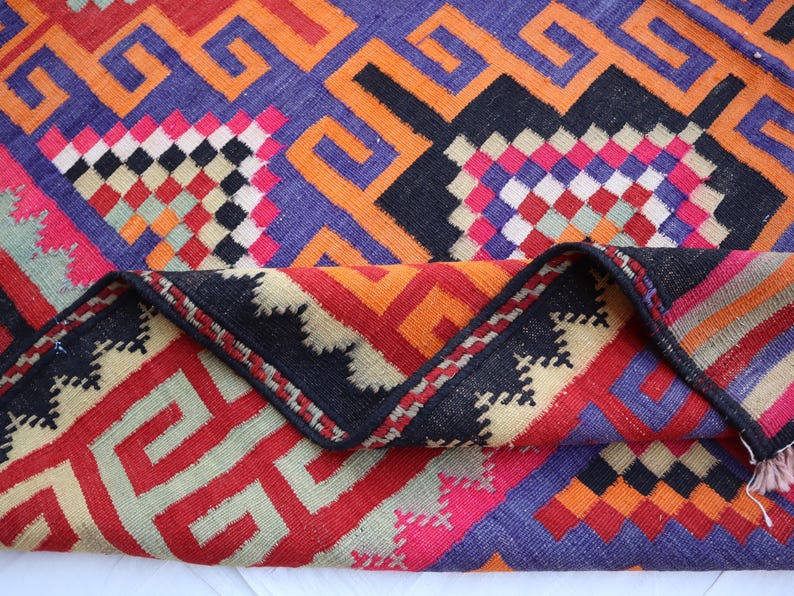 May include: A vibrant, geometric patterned rug with a folded edge. The rug features a mix of colours including red, orange, purple, black, and pink. The design includes repeating geometric shapes and patterns, creating a visually striking textile.