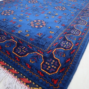 May include: A rectangular area rug featuring a vibrant blue field with repeating geometric floral motifs in red, yellow, and dark blue. The rug has a wide border with intricate patterns and white fringe along the short edge, showcasing a dense pile.