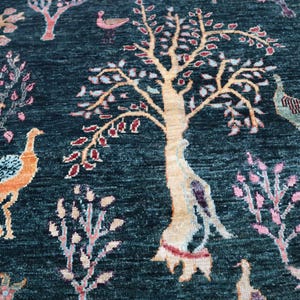 May include: A teal blue rug with a stylized tree design in the center. The tree has a light brown trunk and branches with pink and red leaves. There are also stylized birds and flowers throughout the rug.