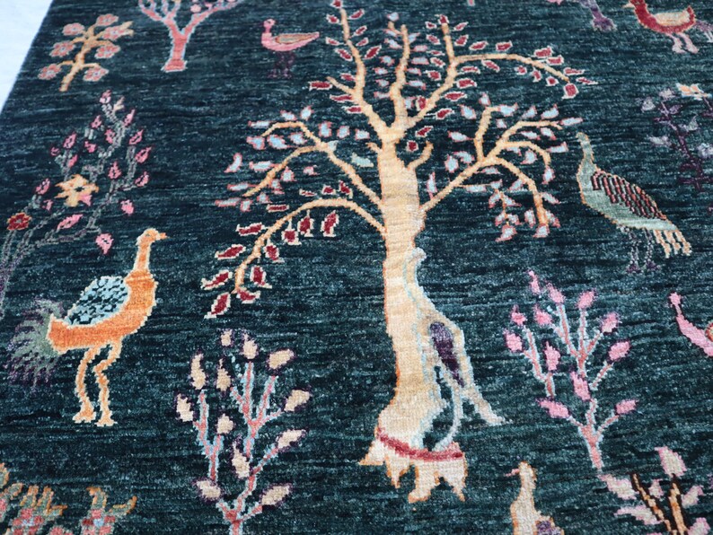 May include: A teal blue rug with a stylized tree design in the center. The tree has a light brown trunk and branches with pink and red leaves. There are also stylized birds and flowers throughout the rug.