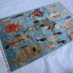 May include: A blue rug with a fringe border features a colorful design of various fish. The rug is decorated with a variety of fish in different colors and patterns, including red, orange, yellow, green, blue, and brown.