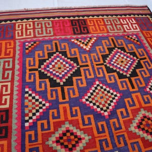 May include: A colourful, geometric patterned rug with a central field of purple, orange, and black. The rug features repeating square and diamond motifs in red, pink, and green. The border is a mix of red, orange, and black.
