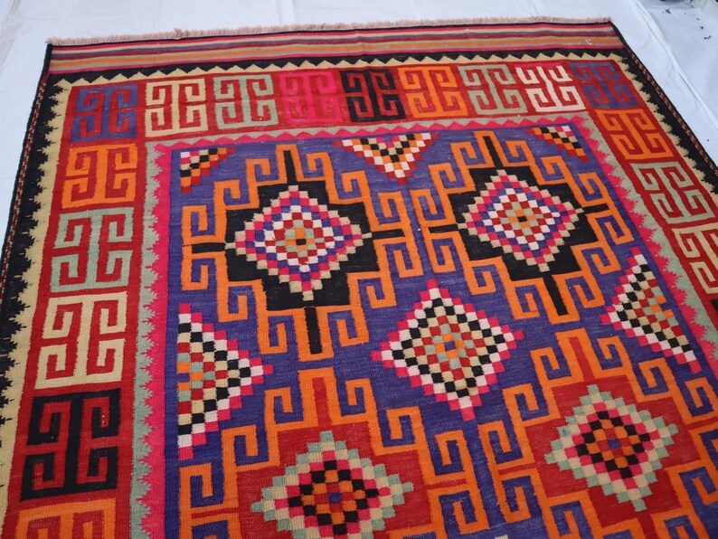 May include: A colourful, geometric patterned rug with a central field of purple, orange, and black. The rug features repeating square and diamond motifs in red, pink, and green. The border is a mix of red, orange, and black.