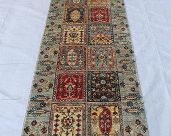 Könnte beinhalten: Ein rechteckiger, gemusterter Teppich mit einer zentralen Säule aus quadratischen Designs in verschiedenen Farben. Der Teppich hat einen hellgrünen Hintergrund mit floralen und geometrischen Mustern und einen Fransenrand.