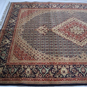 Puede incluir: Una alfombra persa tradicional con fondo azul marino y intrincados patrones florales en tonos rojos, crema y oro. La alfombra tiene un medall&oacute;n central en forma de diamante y un borde con motivos florales repetidos.
