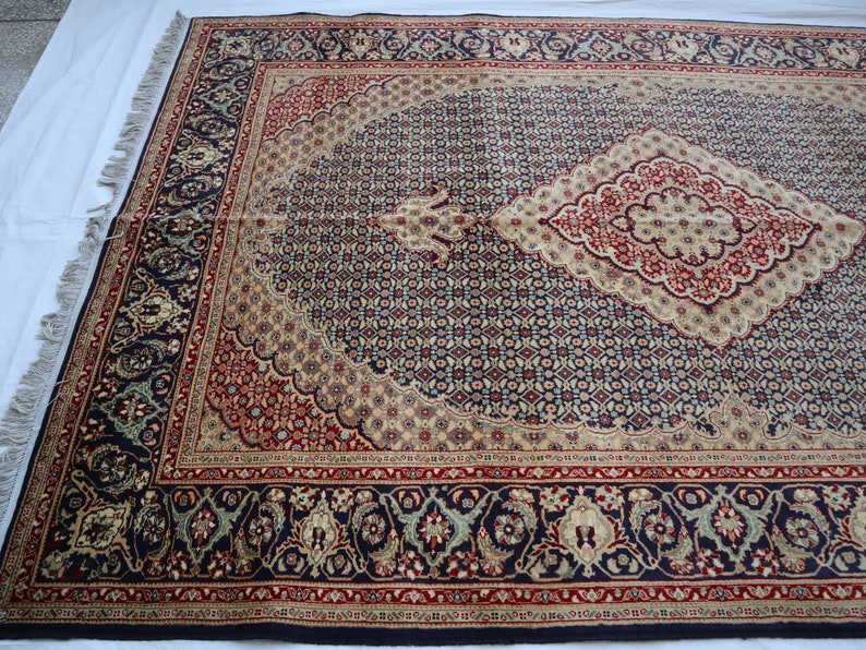 Puede incluir: Una alfombra persa tradicional con fondo azul marino y intrincados patrones florales en tonos rojos, crema y oro. La alfombra tiene un medall&oacute;n central en forma de diamante y un borde con motivos florales repetidos.