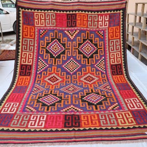 May include: A colourful, rectangular rug with a geometric pattern. The rug features a central design in purple, orange, and black, surrounded by a red border with repeating square motifs. The rug's edges are finished with a fringe.