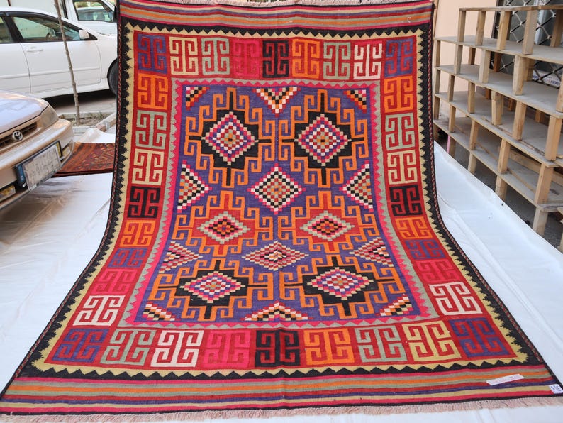 May include: A colourful, rectangular rug with a geometric pattern. The rug features a central design in purple, orange, and black, surrounded by a red border with repeating square motifs. The rug's edges are finished with a fringe.