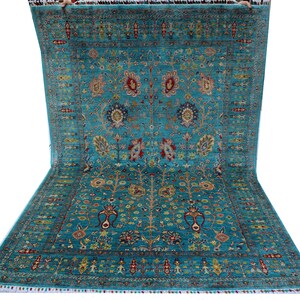 Könnte beinhalten: Ein großer, rechteckiger türkisfarbener Teppich mit einem komplizierten Blumenmuster. Der Teppich hat ein zentrales Medaillon-Design mit roten, gelben und blauen Akzenten. Die Kanten sind mit Fransen versehen.