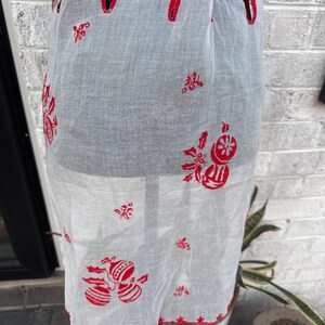 May include: A sheer white apron featuring red embroidered holiday motifs including ornaments, holly leaves, and berries. The apron has a scalloped hem with red stitching, a tie waist, and a repeating pattern of festive decorations across the fabric.