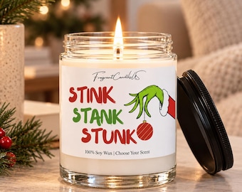 Puede incluir: Una vela en frasco de vidrio transparente con una etiqueta blanca que muestra el texto STINK STANK STUNK en rojo y verde, junto a una mano verde sosteniendo un adorno rojo. La vela tiene una mecha encendida y una tapa negra al lado.