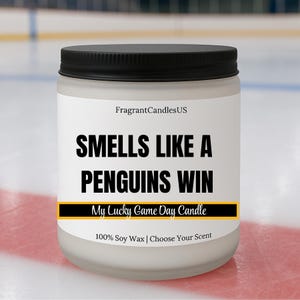 May include: A white jar candle with a black lid. The label reads "SMELLS LIKE A PENGUINS WIN" in bold black letters. Below, it says "My Lucky Game Day Candle". The candle is made of 100% soy wax.