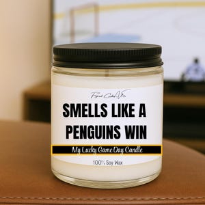 May include: A glass jar candle with a black lid and a white label. The label reads "SMELLS LIKE A PENGUINS WIN" in bold black letters. Below the text is a yellow and black banner that says "My Lucky Game Day Candle".