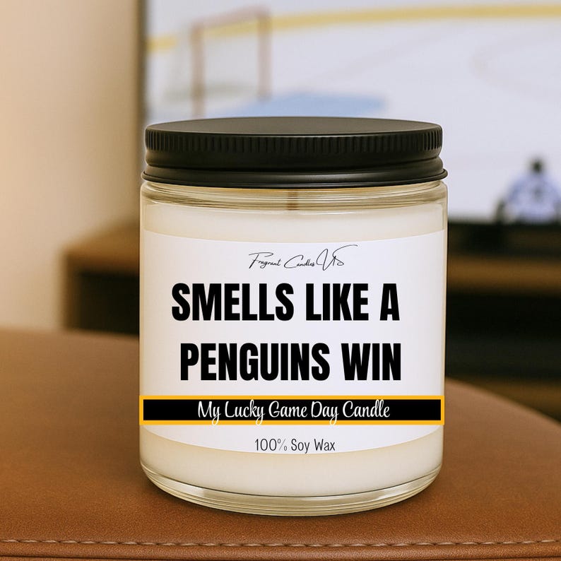 May include: A glass jar candle with a black lid and a white label. The label reads "SMELLS LIKE A PENGUINS WIN" in bold black letters. Below the text is a yellow and black banner that says "My Lucky Game Day Candle".