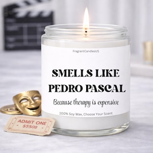 May include: A lit, clear glass jar candle with a white label that reads "SMELLS LIKE PEDRO PASCAL" in black text. Below it says "Because therapy is expensive." The candle is labeled "100% Soy Wax, Choose Your Scent."