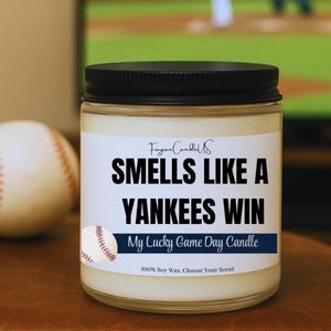 May include: A glass jar candle with a black lid and a white label. The label reads "SMELLS LIKE A YANKEES WIN" with "My Lucky Game Day Candle" below. A baseball graphic is on the label. The candle is 100% soy wax.