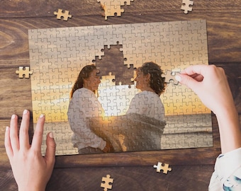 May include: A rectangular jigsaw puzzle featuring a photograph of two people standing together on a beach at sunset. The puzzle is partially assembled, with several loose pieces scattered around the main image on a dark wooden surface.