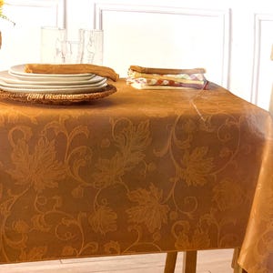 May include: A golden-brown tablecloth with a leaf pattern, set for a meal. White plates, a woven placemat, and napkins are on the table. A wicker vase with yellow flowers and clear glasses are also visible.