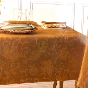 May include: A golden-brown tablecloth with a leaf pattern, set for a meal. White plates, a woven placemat, and napkins are on the table. A wicker vase with yellow flowers and clear glasses are also visible.
