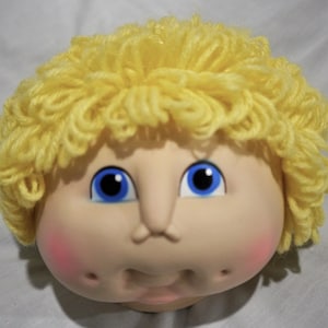 May include: A doll head with yellow yarn hair, blue eyes, and rosy cheeks. The doll has a painted smile and a slightly open mouth.