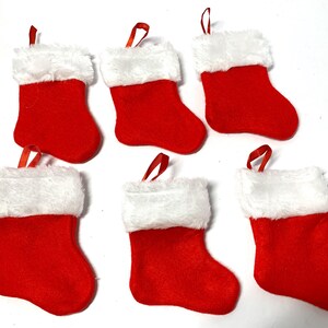 May include: Six miniature Christmas stockings. Each stocking is red with a white, fluffy cuff and a red ribbon loop for hanging. The stockings are arranged on a white surface.
