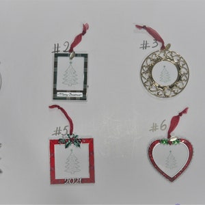 May include: A collection of Christmas tree ornaments in various shapes, including round, square, and heart-shaped. Each ornament features a Christmas tree design and is adorned with a red ribbon for hanging. Some include the year "2021" and holiday accents.