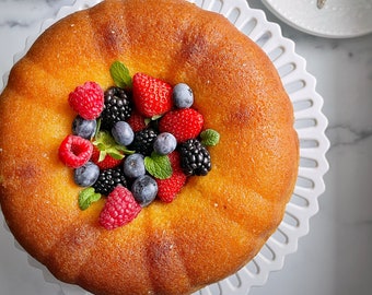 May include: A golden-brown cake, topped with fresh raspberries, blackberries, strawberries, and blueberries, sits on a white, scalloped-edge cake stand. Sprigs of mint add a touch of green. A white plate with silver spoons and a blue-striped napkin are nearby.