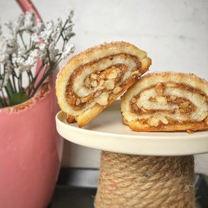May include: Two cinnamon rolls with a walnut filling are displayed on a white cake stand with a rope base. The rolls are on a white plate. The stand is on a black surface.