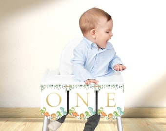 Könnte beinhalten: Ein weißer Hochstuhl mit einem grünen und goldenen Banner, auf dem "ONE" mit einem Dinosaurier-Motiv steht. Ein Baby sitzt im Hochstuhl.