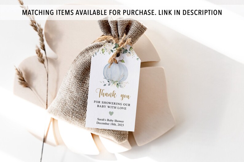May include: A small burlap bag tied with twine, holding a gift tag. The tag features a watercolor illustration of a blue pumpkin with floral accents. Text on the tag reads "Thank you for showering our baby with love." The baby shower date is December 18th, 2023.