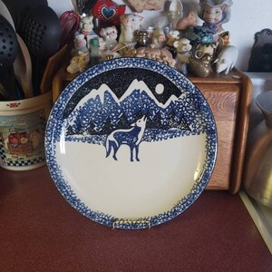 May include: A blue and white ceramic plate with a wolf howling at the moon in a snowy mountain scene. The plate has a speckled blue glaze.