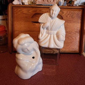 May include: Two white ceramic figurines, one depicting a person holding a fan and the other a person with their arms crossed. Both figurines are in a seated position.