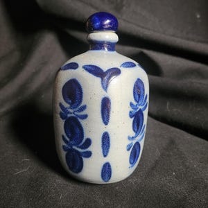 May include: A ceramic bottle with a blue stopper and a light blue body. The body is decorated with dark blue floral patterns. The bottle has a rounded shape and a narrow neck.