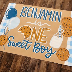 May include: A hand-painted sign with the words "Benjamin is ONE Sweet Boy" in blue and gold lettering. The sign features illustrations of cookies, balloons, and a milk bottle. A blue and white checkered pattern is in the background.