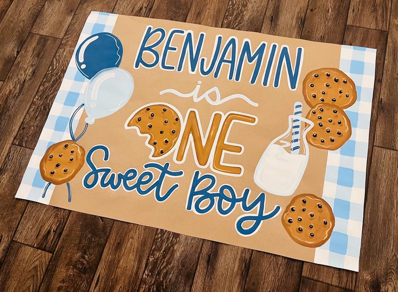 May include: A hand-painted sign with the words "Benjamin is ONE Sweet Boy" in blue and gold lettering. The sign features illustrations of cookies, balloons, and a milk bottle. A blue and white checkered pattern is in the background.