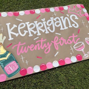 May include: A hand-painted sign on brown paper, celebrating a twenty-first birthday. The sign features the name "Kerrigans" in white, and "twenty first" in pink. It includes a champagne bottle, two wine glasses, and a crown.